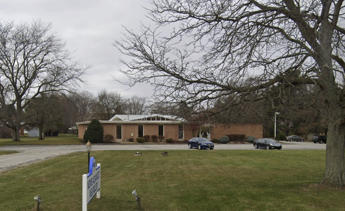 Bellevue Advanced Neurologic clinic, a single-story brick building with a white sign, surrounded by mature trees and a landscaped lawn.